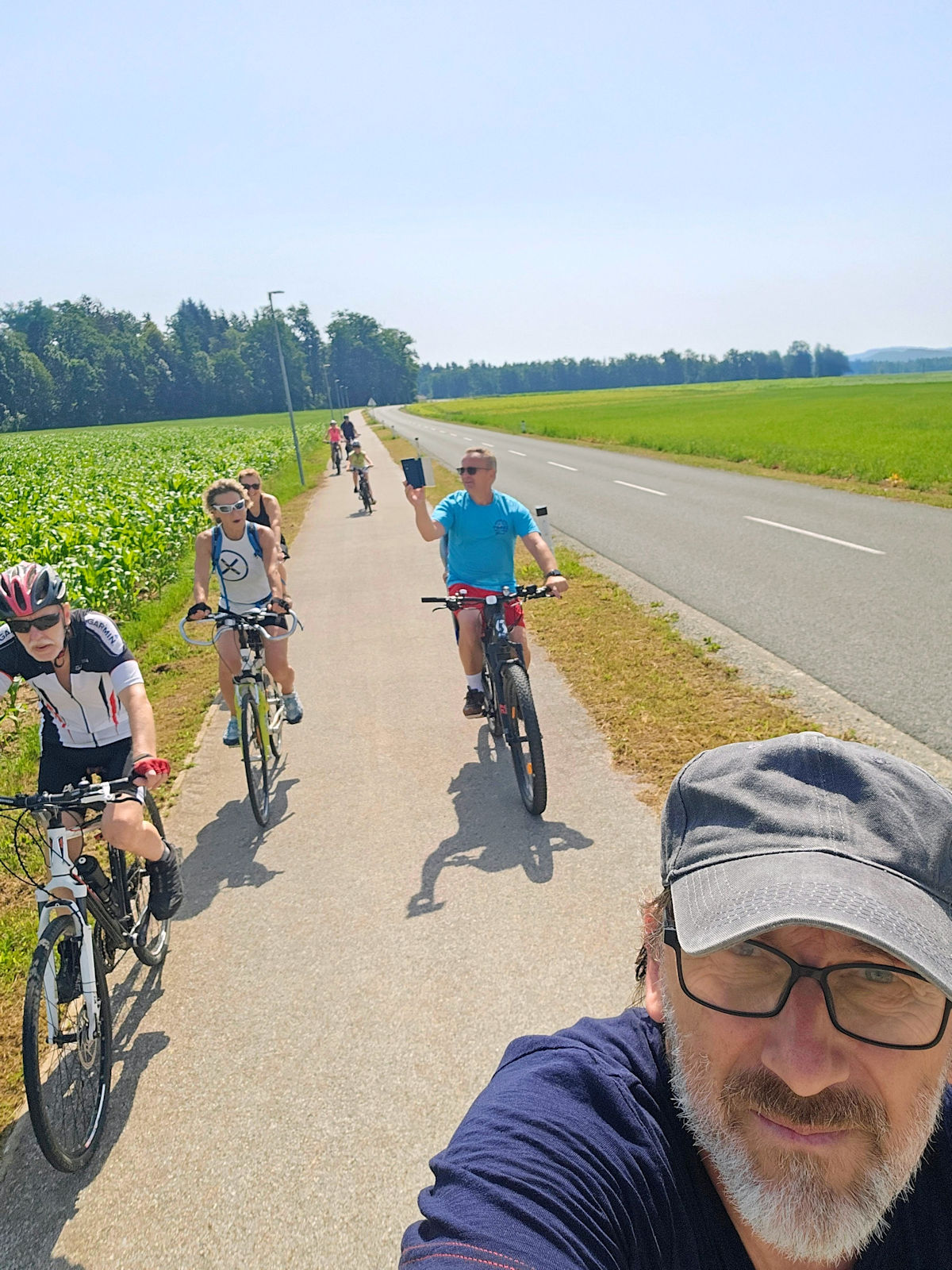 Skupina kolesarjev na urejeni kolesarski poti mimo koruznega polja, v ospredju selfie organizatorja