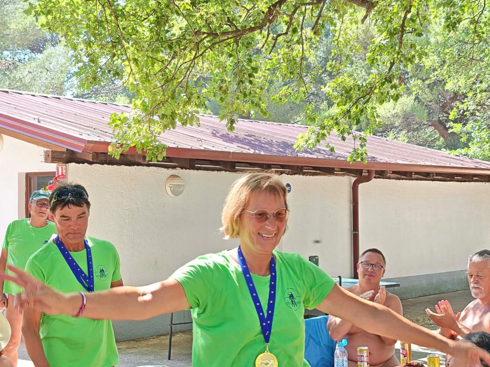 Ženska z medaljo razpre roke od veselja, v ozadju ji drugi ploskajo
