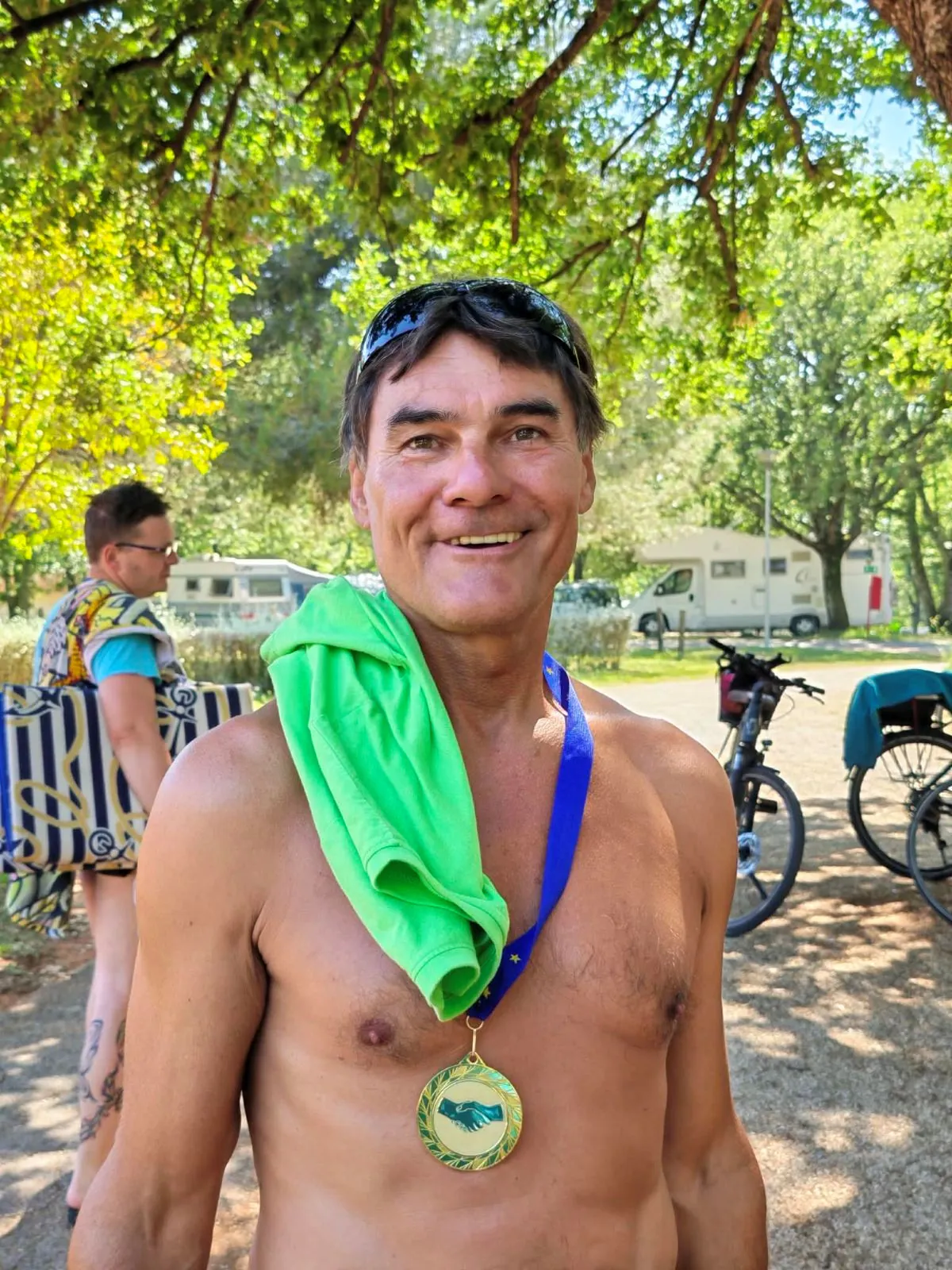 Moški z medaljo in zeleno brisačo čez ramo se nasmehne v kampu