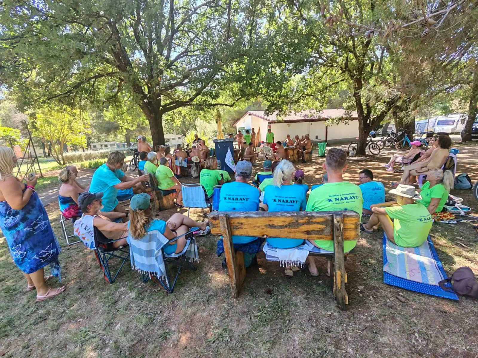 Skupina članov v zelenih majicah sedi na klopeh pod drevesi in posluša