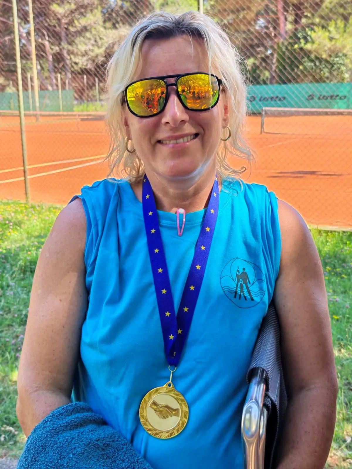 Ženska v zeleni majici kaže zlato medaljo pred teniškim igriščem